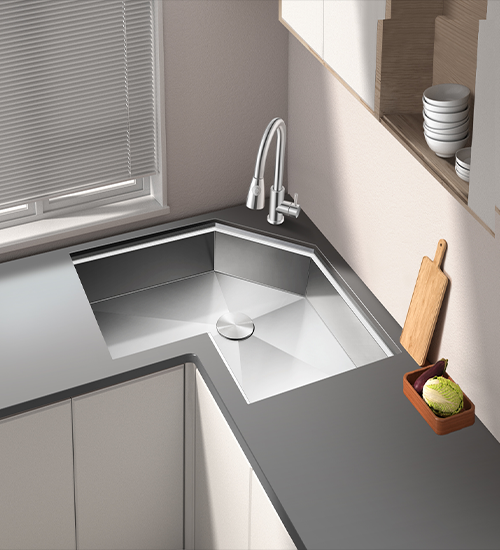 Corner kitchen sink with angled design in stainless steel, embedded in gray countertop with white cabinets, window with blinds, and wooden cutting board visible
